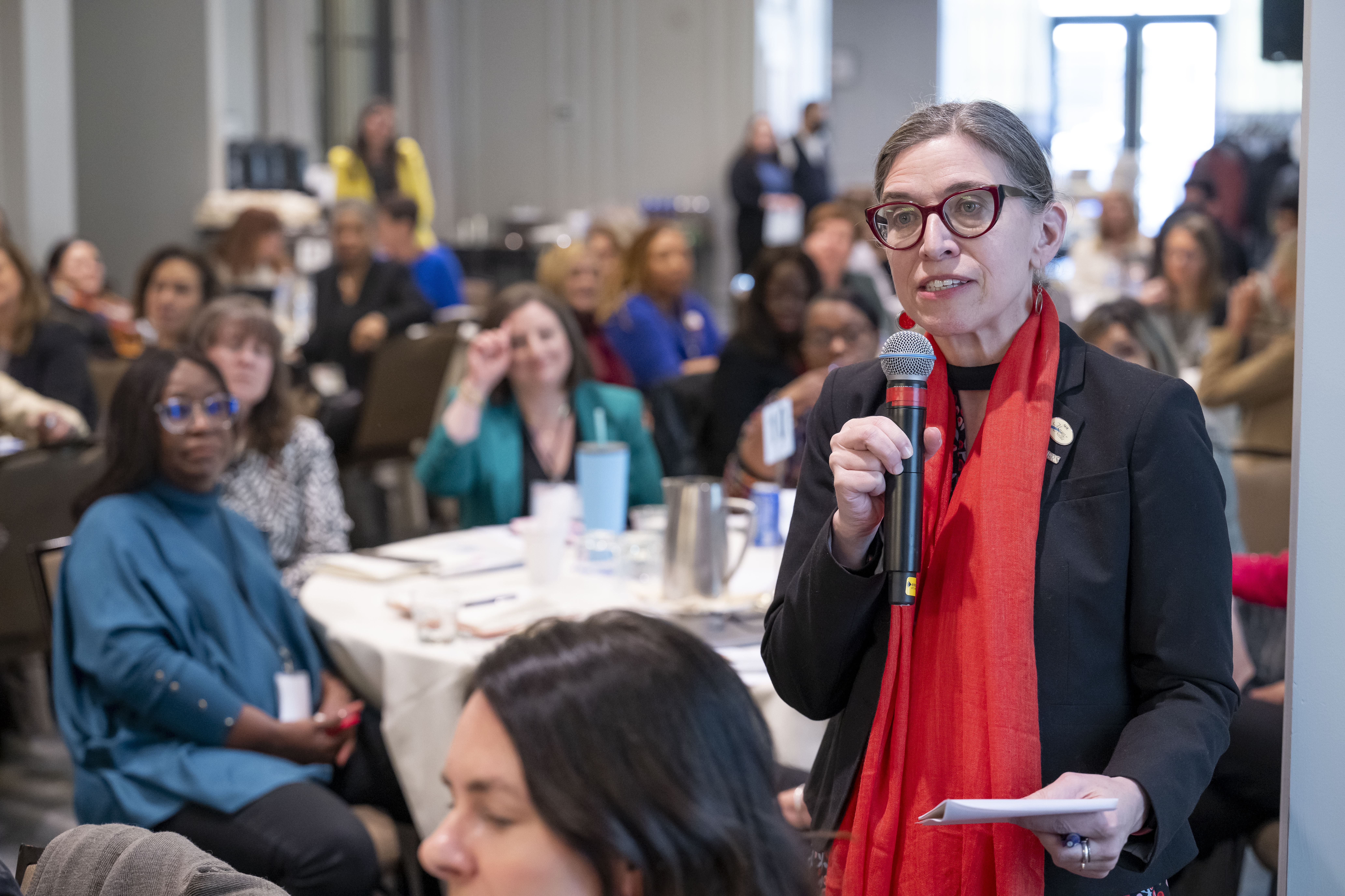 Participants at ACE"s 2024 Women's Conference sitting in the audience of a presentation, with one woman in glasses and a red scarf standing up to ask a question.